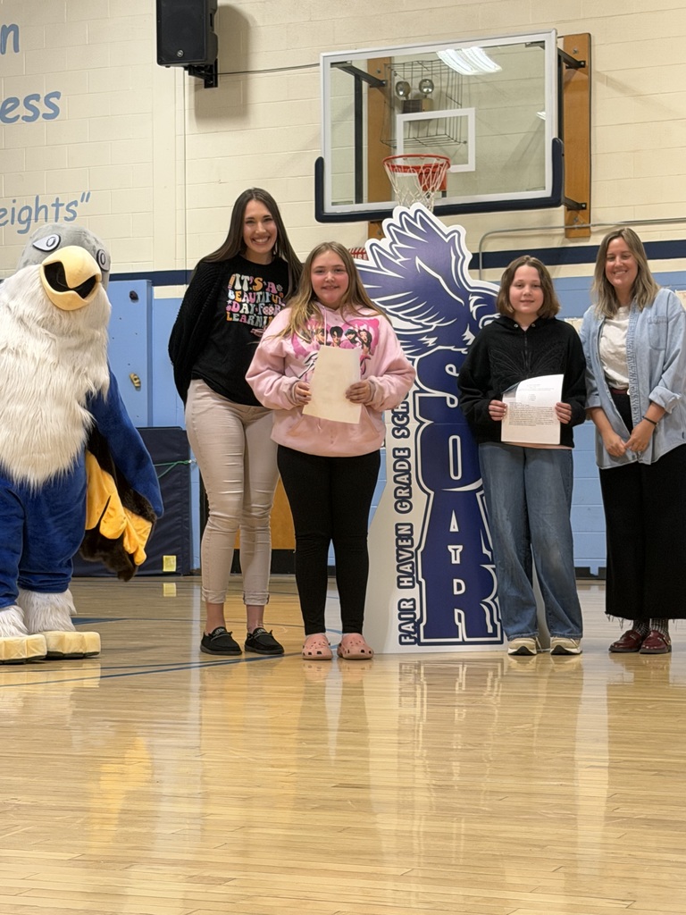 Two students holding certificates and two adults posing with a blue eagle mascot in a school gymnasium in front of a "SOAR" sign.