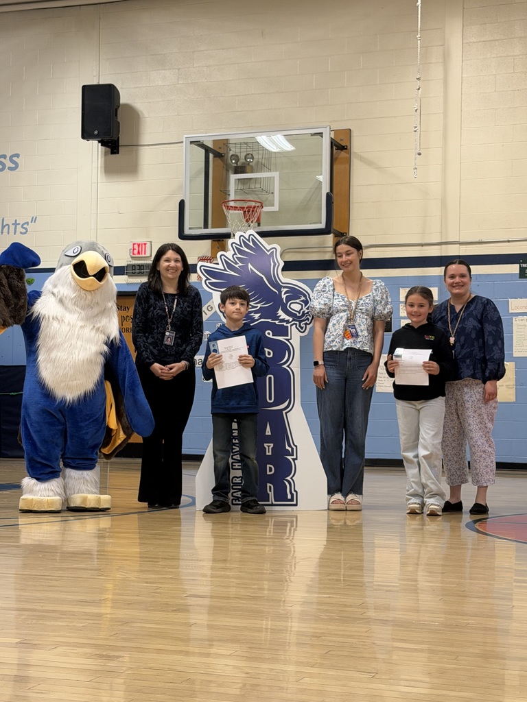 Two students and three adults posing with an eagle mascot in a school gymnasium. The students hold certificates while standing in front of a blue and white "SOAR" cutout and a basketball hoop.