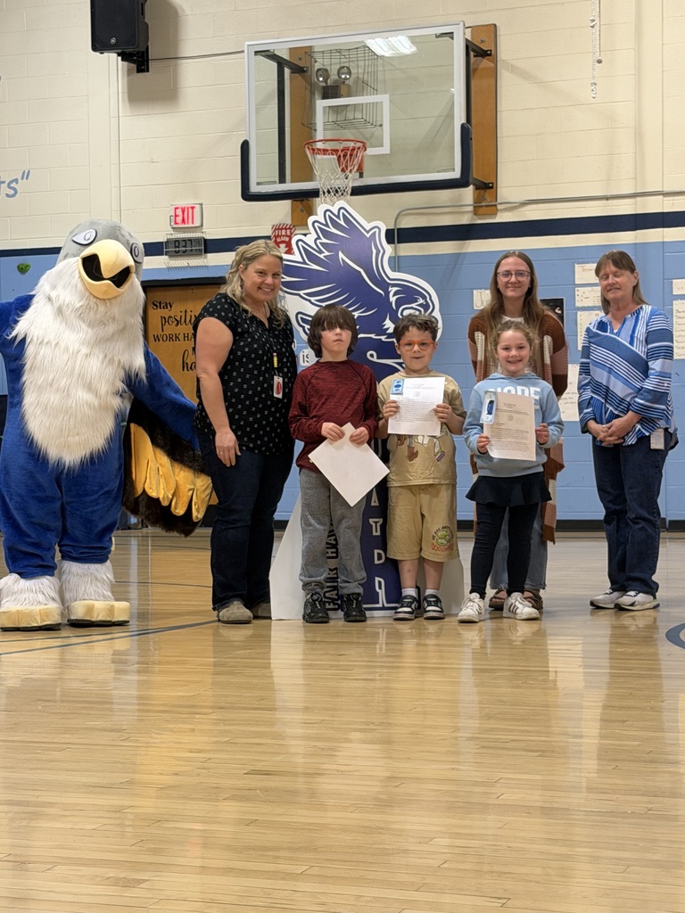 Three students holding certificates stand with three adults and a bird mascot in a school gym, posing in front of a basketball hoop and a large eagle display.