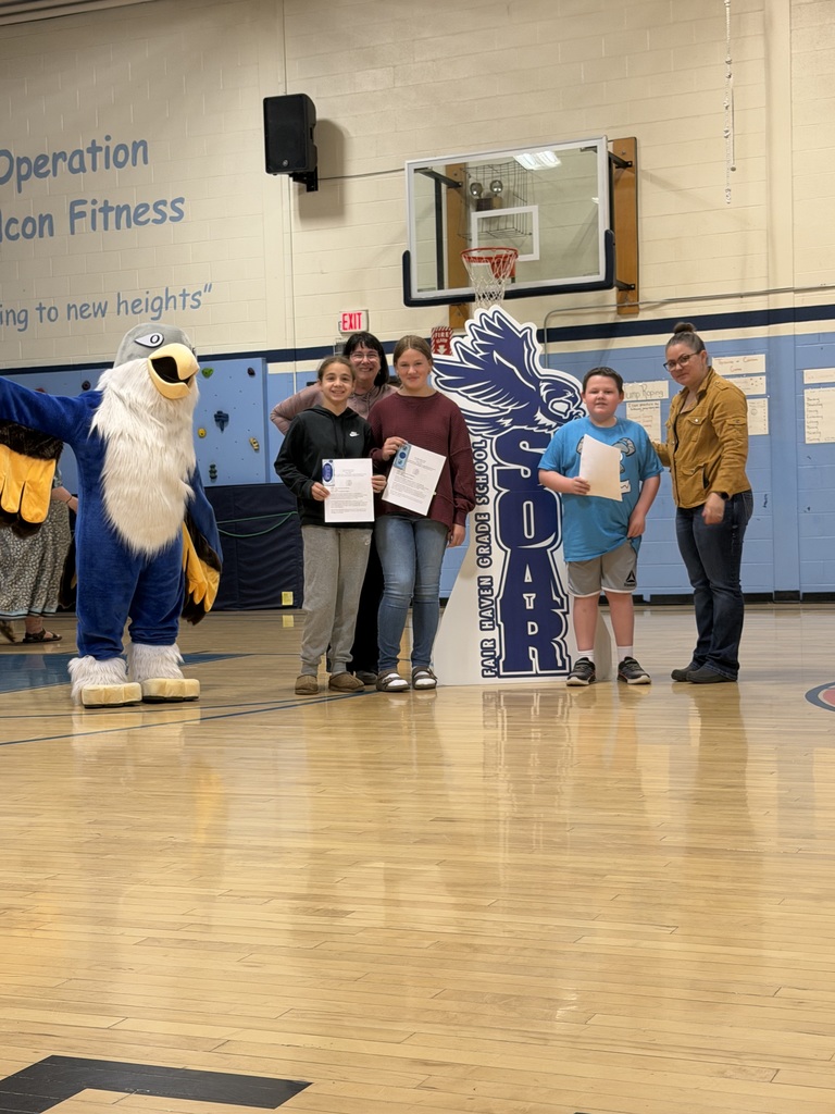A group of four students and two adults stand in a school gymnasium holding certificates, posing next to a large bird mascot and a school eagle sign, with a basketball hoop and gym wall in the background.