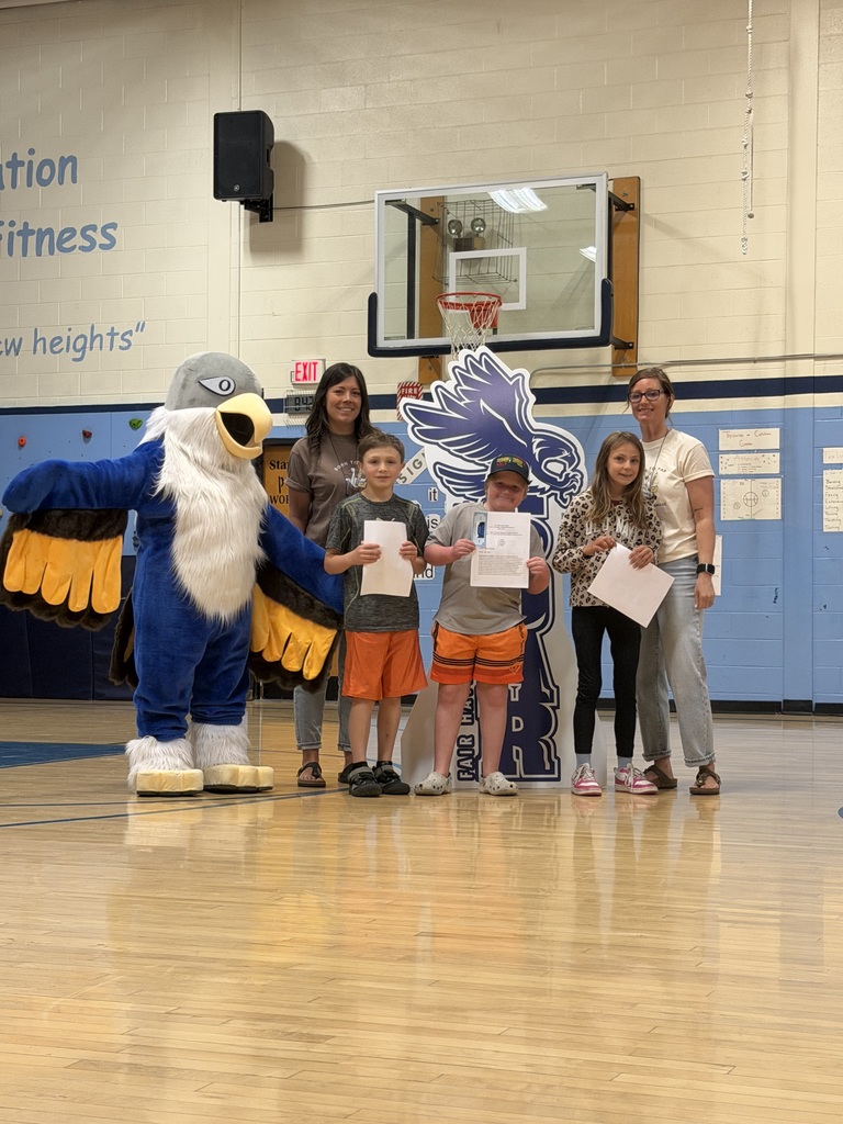 Three students and two adults standing in a school gymnasium next to an eagle mascot, holding certificates in front of a basketball hoop.
