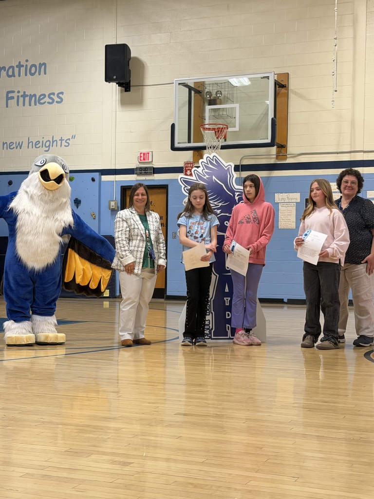 Three students holding certificates and two adults posing with an eagle mascot in a school gymnasium. They are standing in front of a blue "SOAR" sign and a basketball hoop.