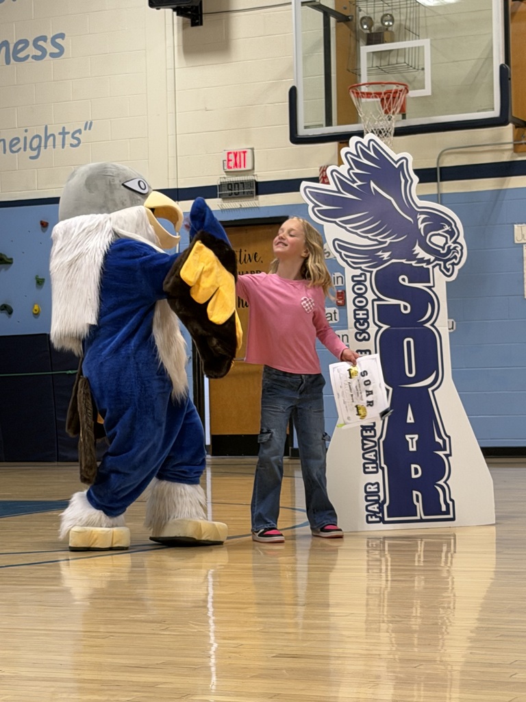 A student in a pink shirt high-fives an eagle mascot in a school gymnasium while holding a "SOAR" certificate.