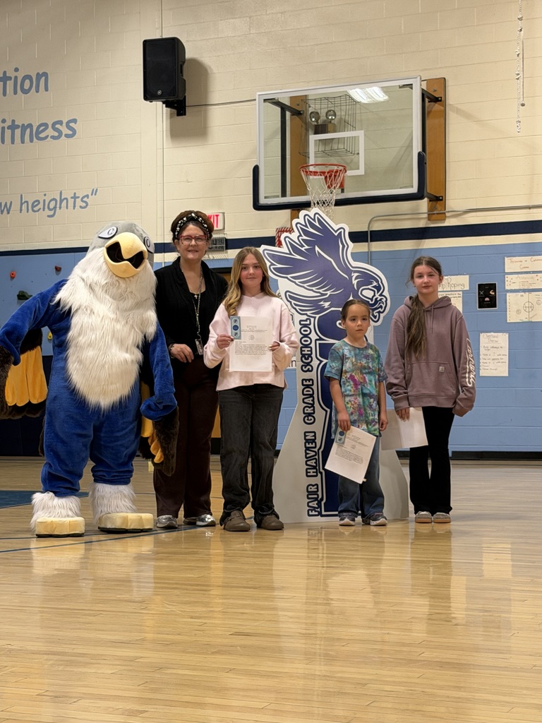 Three students holding certificates and one adult posing with a blue eagle mascot in a school gymnasium in front of a "Fair Haven Grade School" sign.