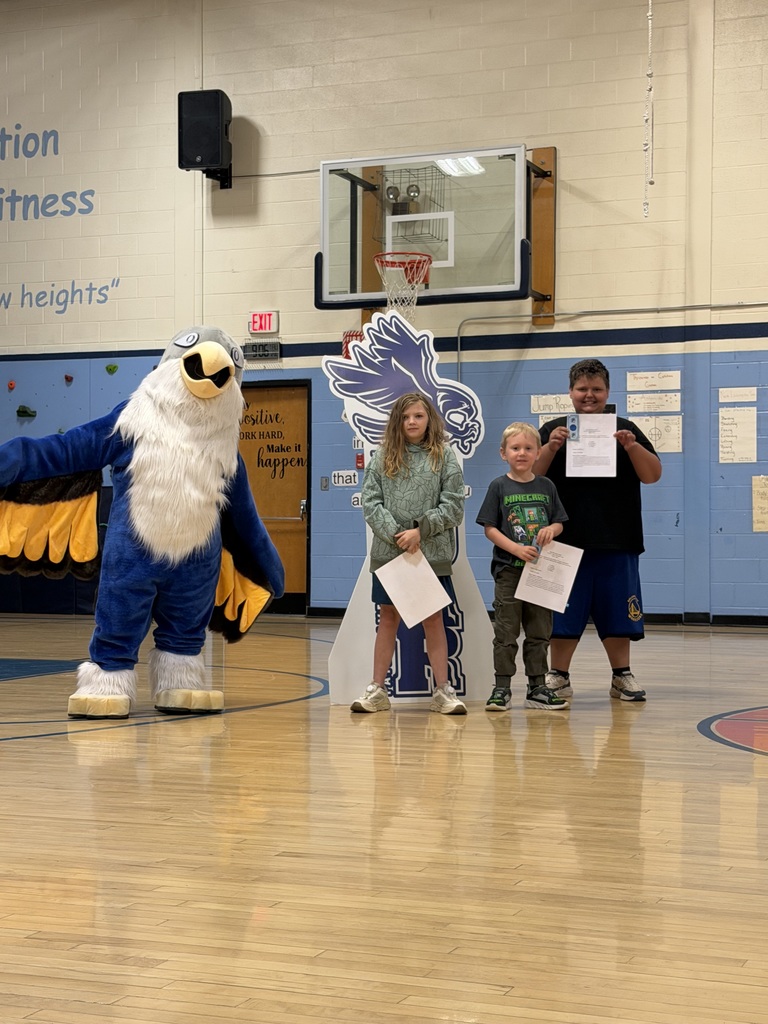 Three students holding certificates pose with a blue and white eagle mascot in a gymnasium in front of a "SOAR" sign.