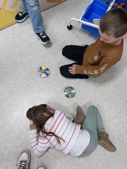 A high-angle close-up of a child kneeling on a speckled classroom floor, pointing their finger toward a spinning top made from a recycled CD and a green bottle cap.