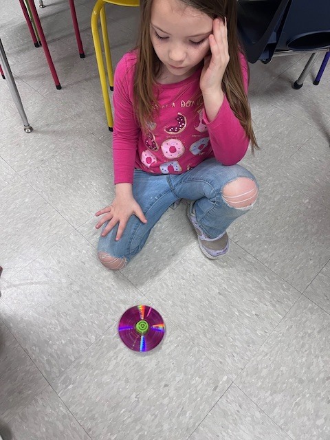 A young girl in a pink doughnut-patterned shirt and ripped jeans crouches on the floor, watching a spinning CD top with a focused expression.