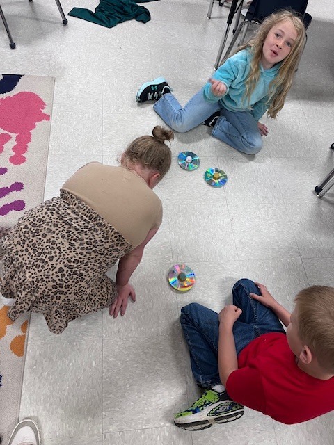 An overhead view of a classroom floor where two children are observing two spinning CD tops. A blue toy bin and the edge of a colorful rug are visible in the background.
