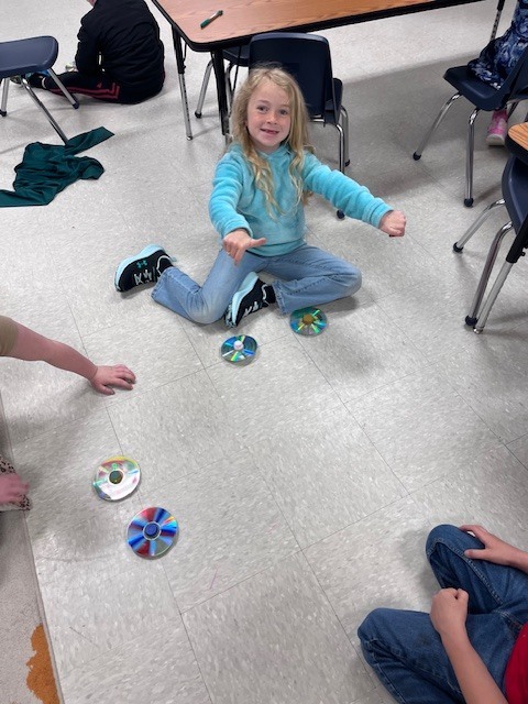 : A young girl with long blonde hair sits cross-legged on a classroom floor, smiling and holding her thumbs up while two CD tops spin in front of her.