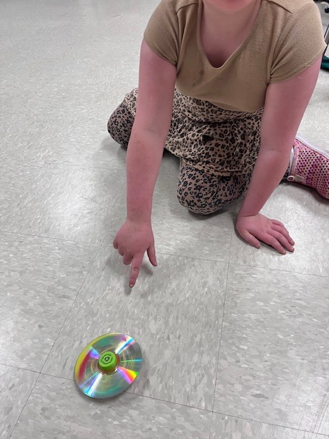 A high-angle close-up of a child kneeling on a speckled classroom floor, pointing their finger toward a spinning top made from a recycled CD and a green bottle cap.
