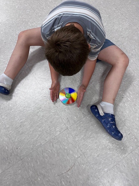 A top-down view of a boy in a striped shirt sitting with his legs spread on the floor, cupping his hands around a rapidly spinning, multicolored CD top to keep it in place.
