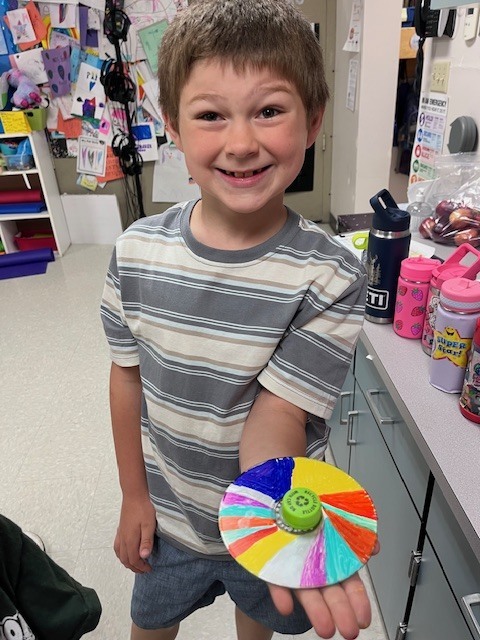 A smiling young boy proudly holds up a handmade spinning top. The top is made from a CD decorated with colorful marker segments and a green bottle cap glued to the center.
