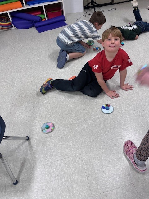 A wide shot of a classroom floor where several children are playing with DIY spinning tops. One boy in a red shirt sits in the foreground looking at the camera, while another boy in a striped shirt experiments with a top in the background.