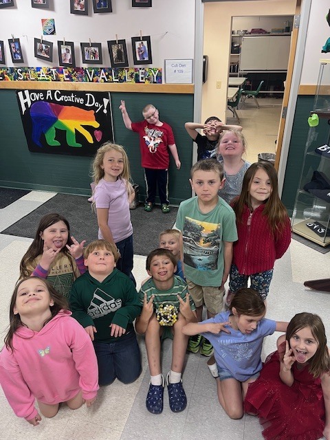 A group of about twelve elementary-aged children pose for a fun group photo in a classroom hallway. Some children are making silly faces and peace signs in front of a "Have a Creative Day!" poster featuring a rainbow bear.