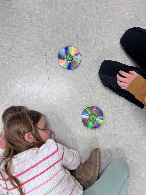 A high-angle shot of two children on the floor watching two CD tops spin. One child is lying down to get a side-view of the motion, while the other sits cross-legged.