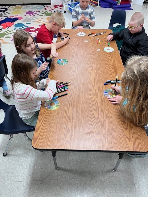 Five children sit around a long wooden table, using colorful permanent markers to draw designs on the reflective surface of CDs as part of a craft project.