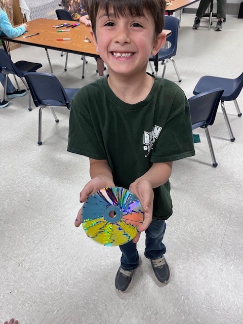A top-down view of a boy in a striped shirt sitting with his legs spread on the floor, cupping his hands around a rapidly spinning, multicolored CD top to keep it in place.