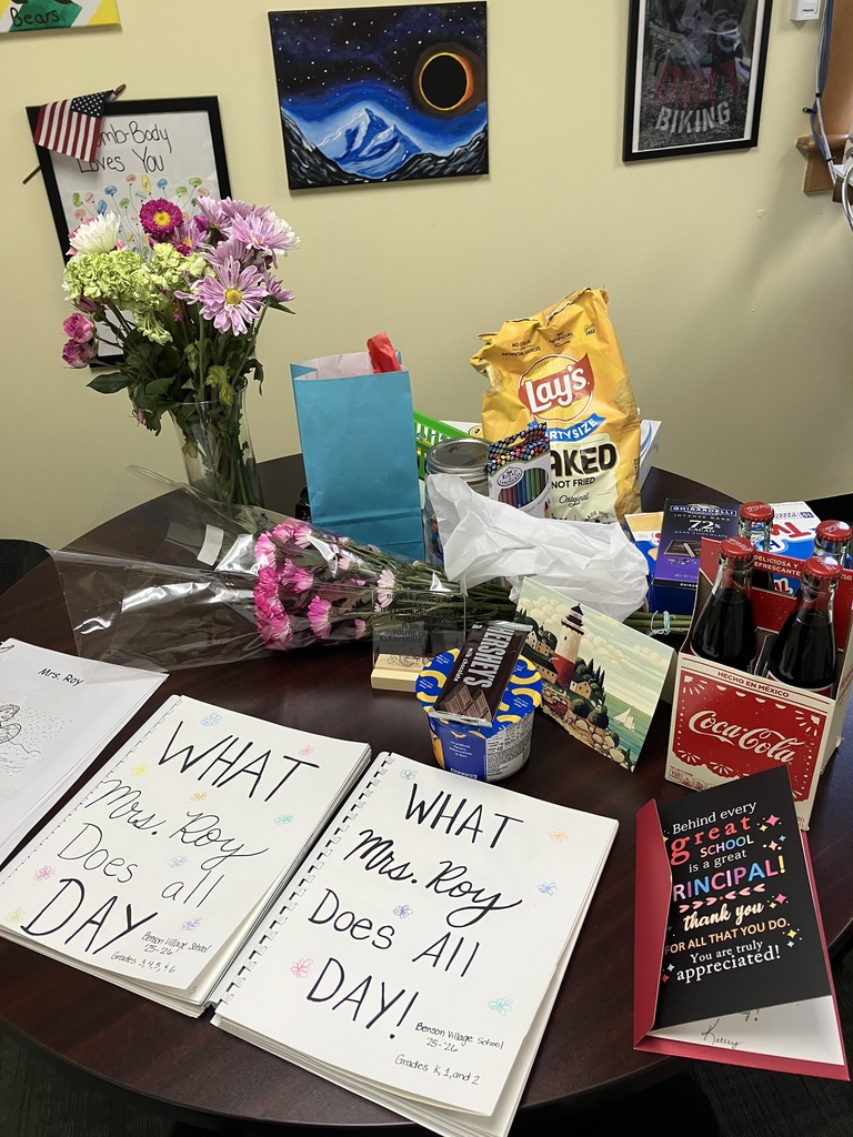 A wooden office table covered in gifts for a principal named Mrs. Roy. Items include several bouquets of pink and white flowers, a glass jar of M&Ms, a 6-pack of Coca-Cola, a Hershey bar, and a cup of mac and cheese. In the foreground are three student-made books titled "WHAT Mrs. Roy Does all DAY!" organized by grade levels.  