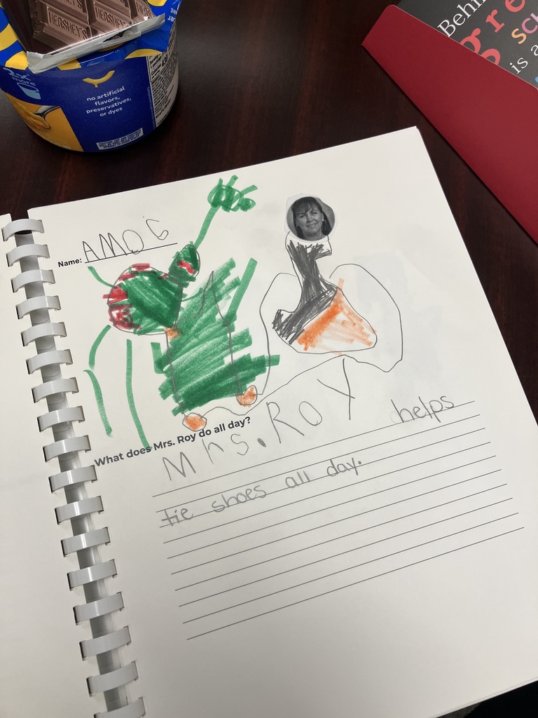 A child’s drawing in a spiral-bound book. The student, named Amos, drew a green figure and a figure with a small cutout photo of Mrs. Roy’s face attached to it. Below the drawing, the student wrote: "Mrs. Roy helps tie shoes all day."