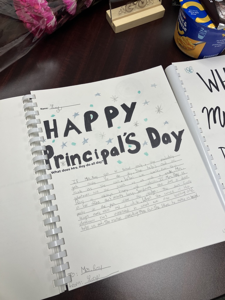  A close-up of a spiral-bound book page titled "HAPPY Principal’s Day" in large, hand-drawn black letters decorated with blue stars. Under the prompt "What does Mrs. Roy do all day?", a student named Lucy has written a heartfelt paragraph in pencil about how the principal gets to school early, greets students with a smile, and "makes everything more fun."  