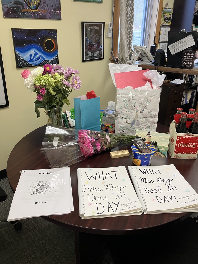  A wooden office table covered in gifts for a principal named Mrs. Roy. Items include several bouquets of pink and white flowers, a glass jar of M&Ms, a 6-pack of Coca-Cola, a Hershey bar, and a cup of mac and cheese. In the foreground are three student-made books titled "WHAT Mrs. Roy Does all DAY!" organized by grade levels. 