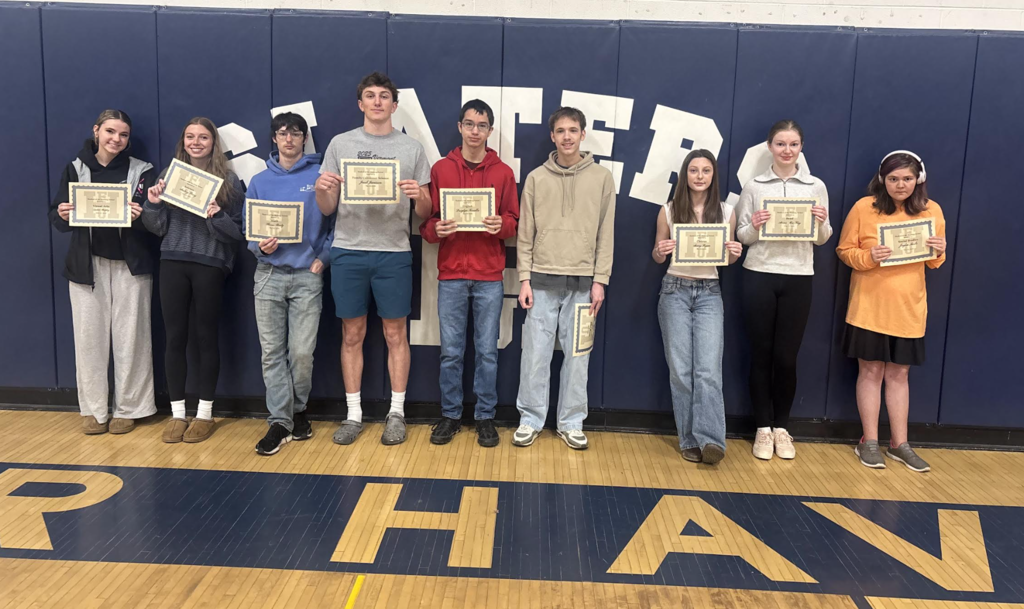 Group of high school students stand in a gym in front of a blue wall with large “Slaters” lettering, each holding a certificate recognizing them as Students of the Marking Period.