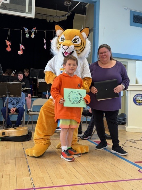 A student in an orange hoodie holds a blue certificate beside a tiger mascot and an adult in a gymnasium with a band seated in the background.