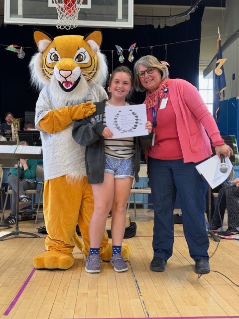 A student holds a white certificate while posing with a tiger mascot and an adult holding a microphone in a school gym.