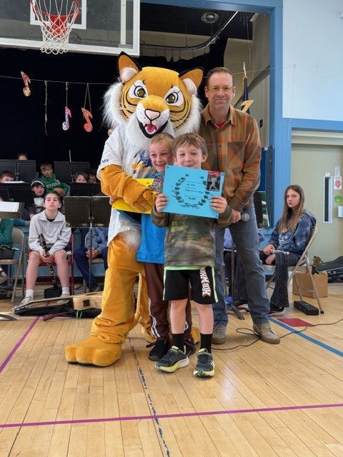 Two elementary students stand with a tiger mascot and an adult in a gym, holding a blue award certificate while a school band sits in the background.