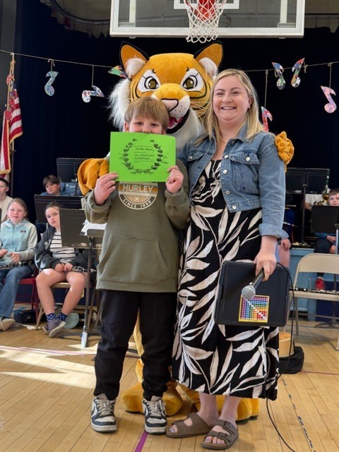 A student holds a green award certificate while standing with a smiling adult and a tiger mascot in a decorated school gym.