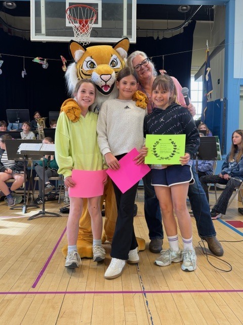 Three students stand together holding certificates with a tiger mascot and an adult in a gymnasium during an award presentation.