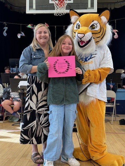A student holds a pink certificate while standing between a tiger mascot and an adult in a gym with musical decorations and students seated behind them.