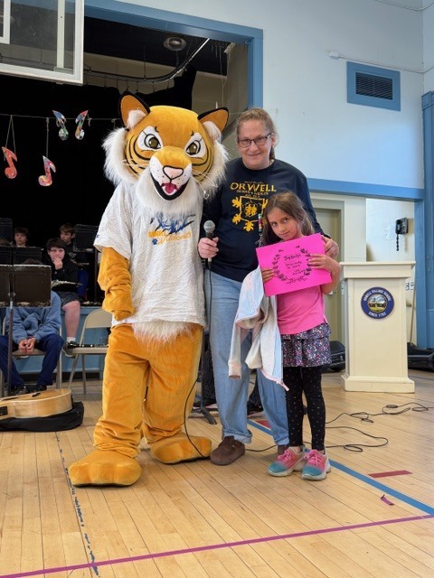 A student holds a pink award certificate while standing with a tiger mascot and an adult holding a microphone in a school gym with musicians behind them.