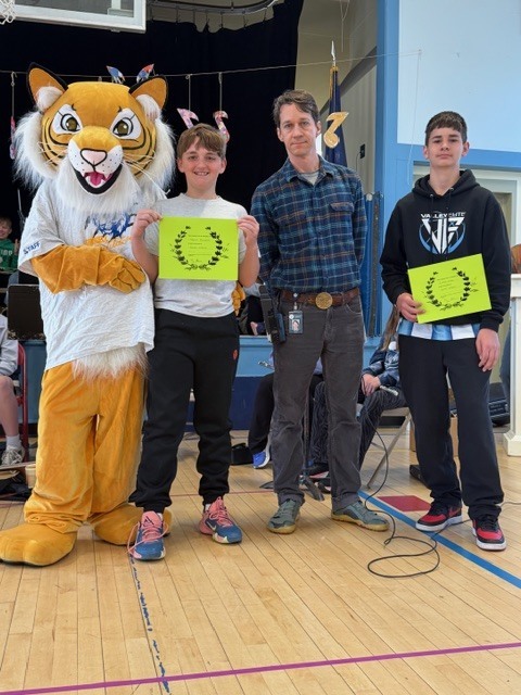 Two students hold bright certificates while standing with a tiger mascot and an adult during an award presentation in a gym with a band in the background.