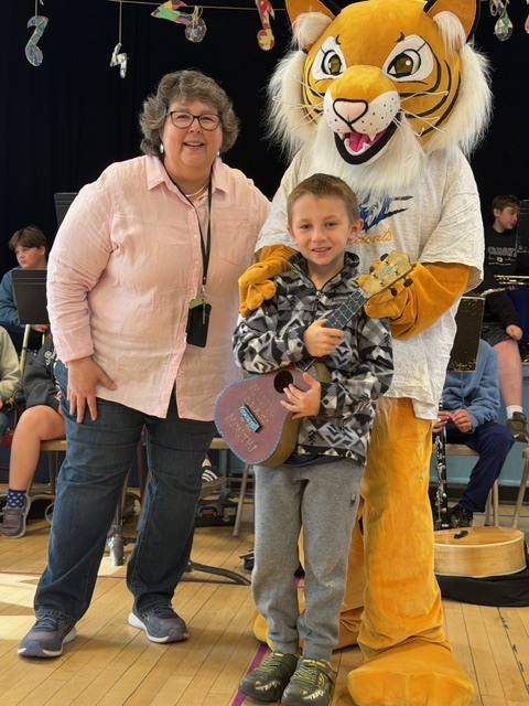 A student holding a small ukulele stands between a teacher and a tiger mascot in a gymnasium, with a school band seated in the background.