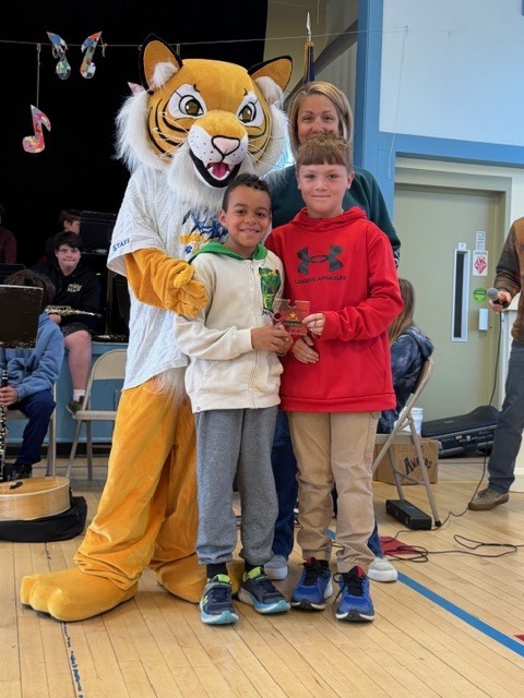 Two elementary students stand together holding a small award while smiling beside a tiger mascot and an adult in a school gym, with a band seated in the background.