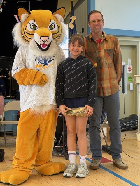 A student smiles while holding a small award, standing next to a tiger mascot and an adult in a school gym during an assembly.