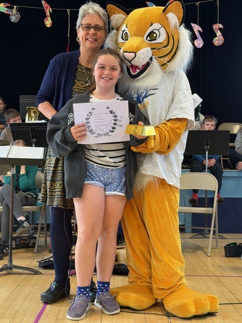 A student holds a certificate and a small award while posing with a teacher and a tiger mascot during a school assembly in a gym with musicians behind them.
