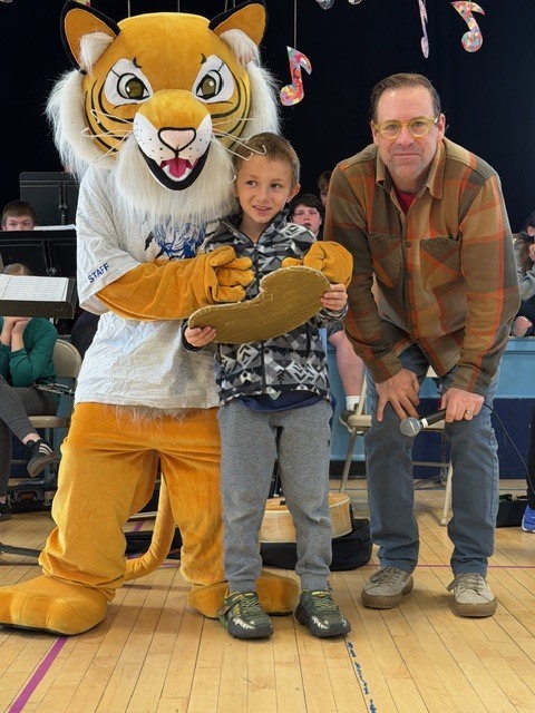 A student holds a unique award while standing beside a tiger mascot and an adult holding a microphone, with a band and musical decorations visible in the background.