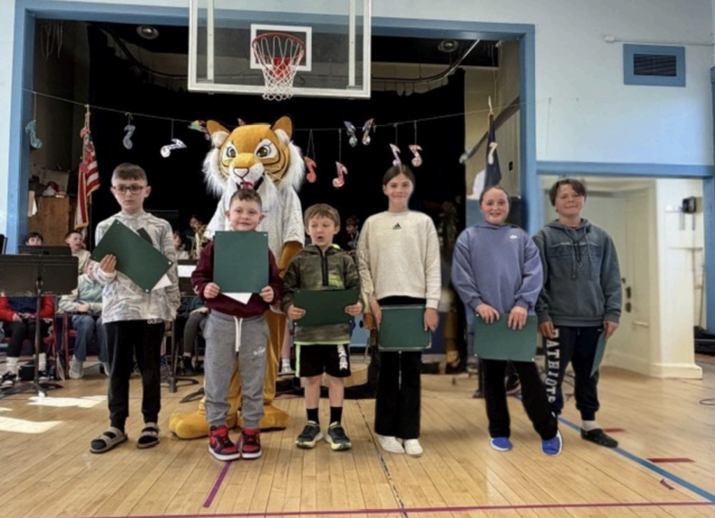 A group of elementary students stand in a line in a school gym holding green certificates, with a tiger mascot behind them. A stage with seated band students and hanging music note decorations is visible in the background under a basketball hoop.