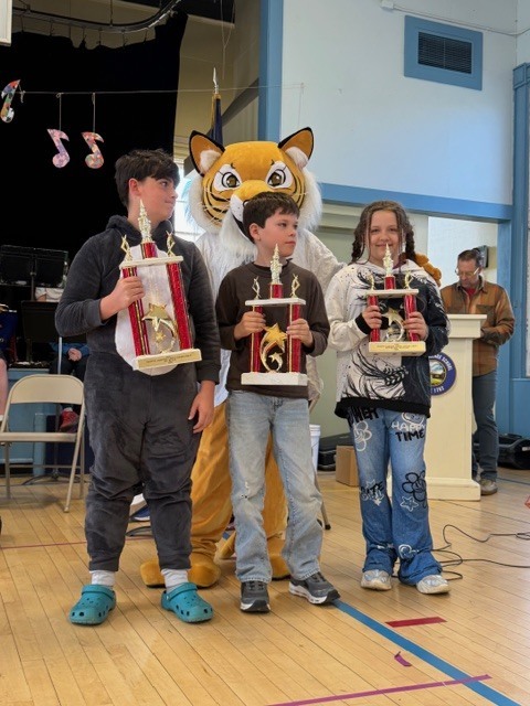 Three students hold trophies while standing together in a gymnasium with a tiger mascot behind them; a staff member stands at a podium in the background during an assembly.