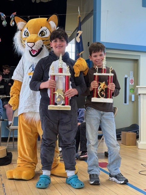 Two students stand in a school gym smiling and holding large trophies, with a tiger mascot behind them. Musical decorations and seated band students are visible in the background.