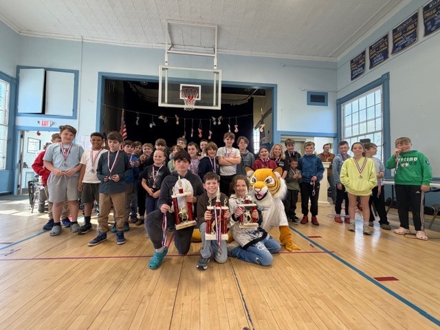 A large group of students gathers in a school gym for a group photo, many wearing medals, while three students kneel in front holding trophies and a tiger mascot crouches beside them near the stage.