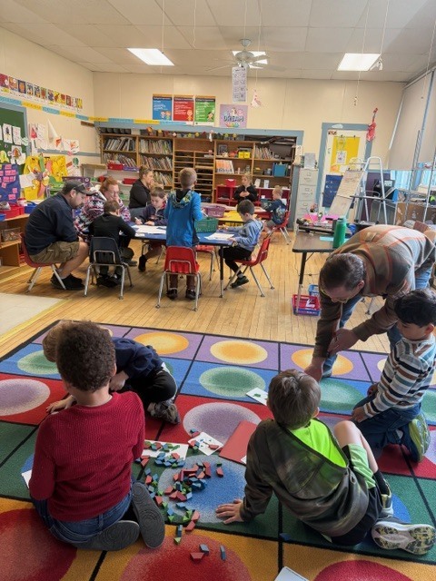 Students and adults work together in a colorful elementary classroom, with some children building on a rug with blocks while others sit at tables completing activities.