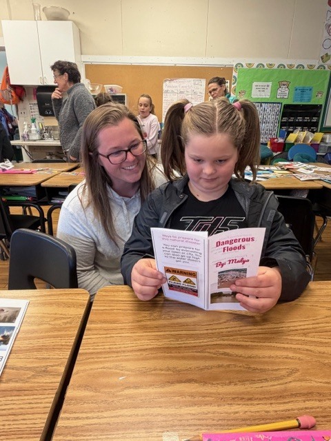 A student reads a small booklet titled “Dangerous Floods” while an adult sits beside them smiling in a classroom setting.