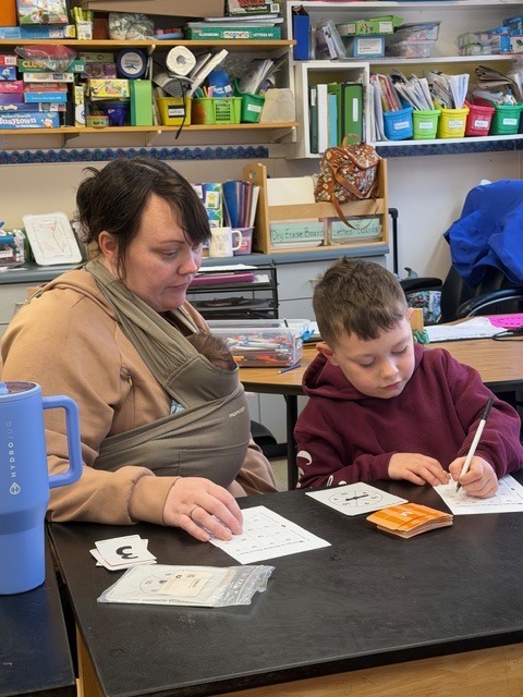 A young student writes on a worksheet while an adult sits beside them for support at a classroom table with learning materials.