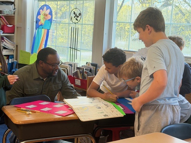 A teacher sits with a small group of students around a desk, guiding them as they look at a large book and complete an activity together.