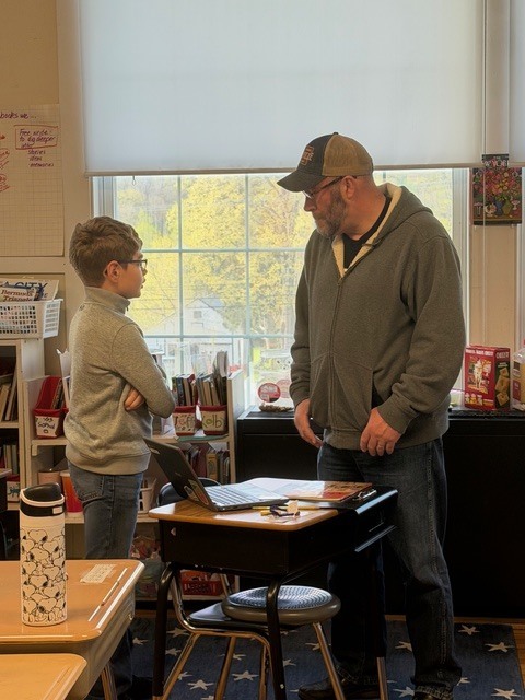A student stands beside an adult at a desk, discussing work on a laptop in a classroom with a large window behind them.