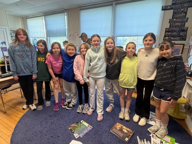 A group of students stand together on a classroom rug, smiling for a group photo with books and classroom materials scattered nearby.