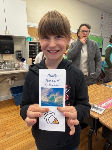 A smiling student holds up a booklet titled “Deadly Tsunamis” in a classroom while a teacher stands in the background.